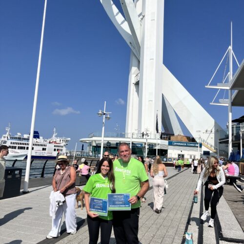 father and daughter in green t shirts holding a medal and certificate stood in the sun infront of white spinnaker tower