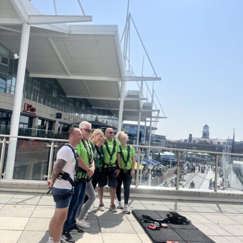 group of people stood to the side of the camera wearing green t shirts and climbing harnessess on a platfrom in shopping area at gunwharf quays