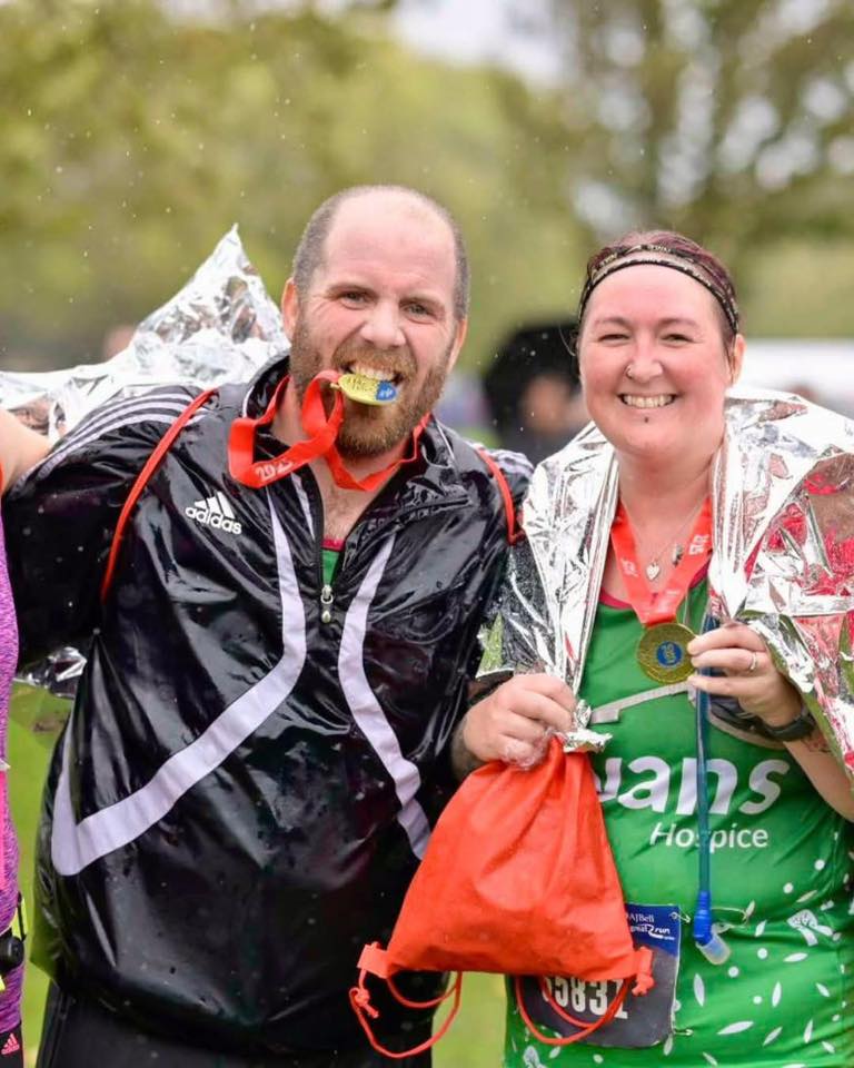 A bald man biting on a gold medal with a red lanyard, wearing a black adidas jacket, a woman with red hair, wearing a green Rowans Hospice vest, with a foil wrap over her and holding a gold medal