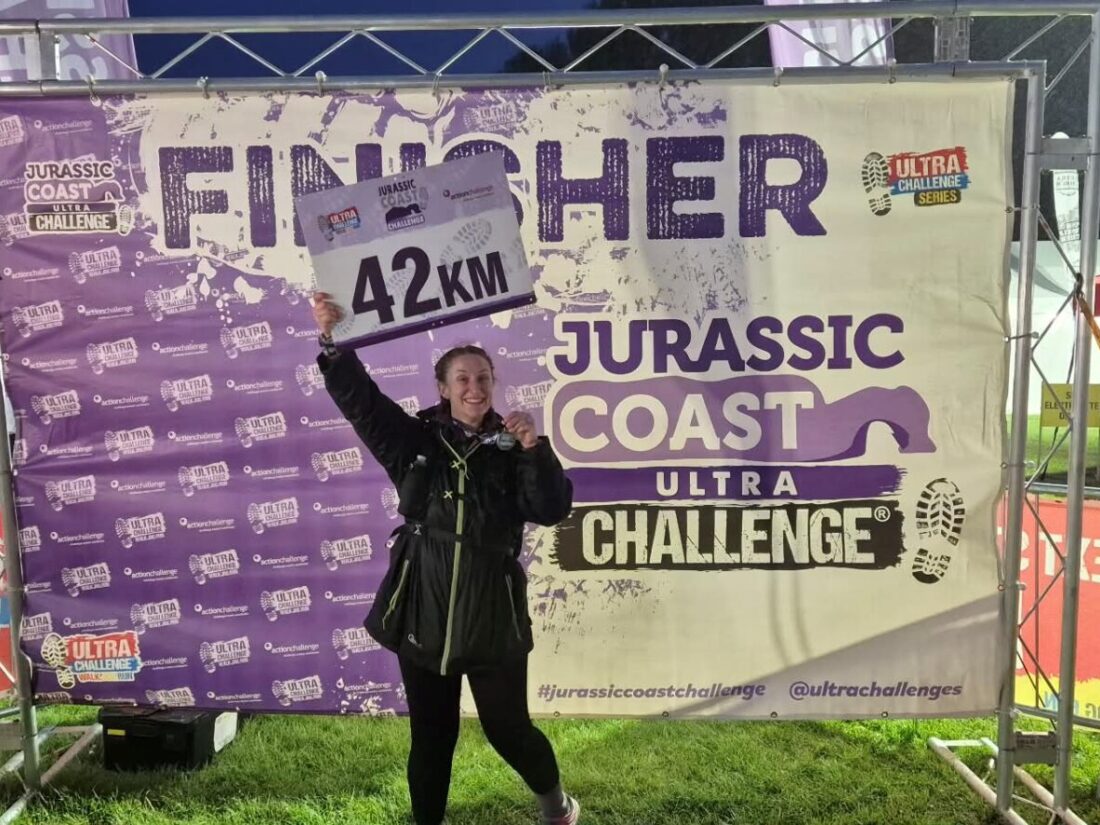 Woman wearing all black holding a sign that says "42KM" behind her is a banner that says "FINISHER, Jurassic Coast Ultra Challenge"