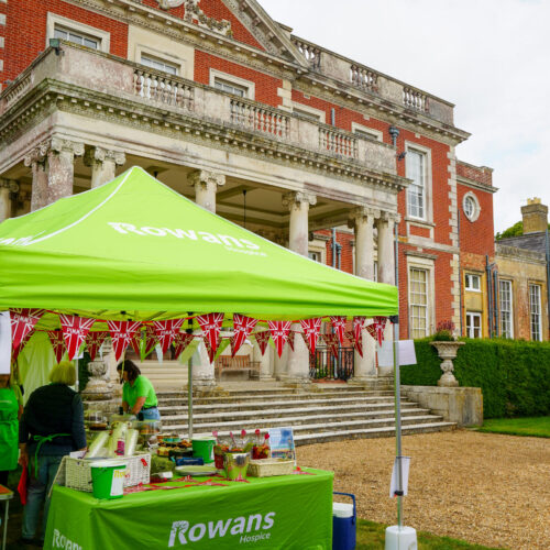 Gazebo with green roof with "Rowans Hospice" logo, green table with green table cloth saying "Rowans Hospice", in front of an Edwardian mansion.
