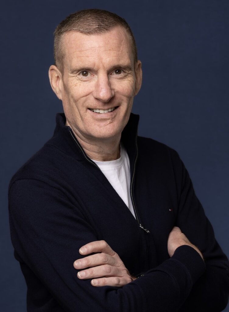 Picture of man from middle of the torso up, the man has his arms crossed and looks at the camera at three quarters, he is smiling. The man has short dark hair and wears a black v neck jumper with a white shirt underneath