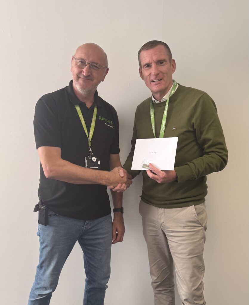 A man in a black polo shirt and jeans is shaking hands with another man wearing a green sweater and beige trousers. Both are smiling and wearing green lanyards. The man in the green sweater is holding an envelope and a small gift. They are standing in front of a plain light-colored wall.