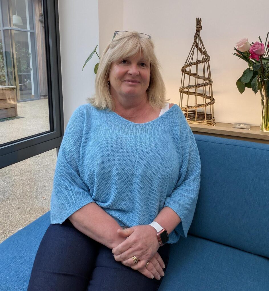 Middle aged woman, short blond hair, sat on a blue sofa, wearing a light blue sparkly jumper and dark blue jeans, her hands are crossed over each other