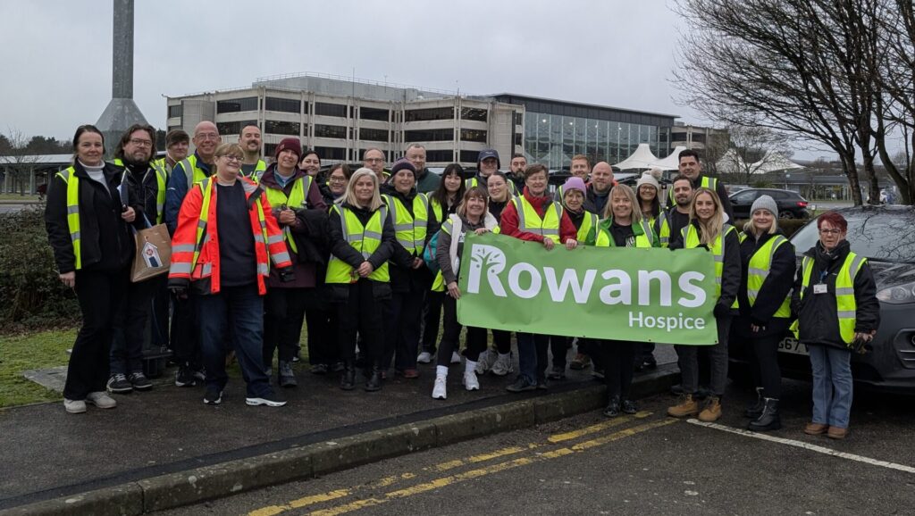 A large group of volunteers wearing yellow hi-vis pose together in a parking lot, holding a large green banner that reads "Rowans Hospice." A modern office building and overcast winter sky are in the background.