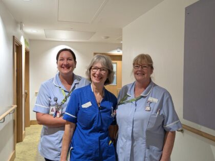 Three ladies posing and smiling in a corridor. The lady on the left is a healthcare support worker, she is wearing light blue scrubs and her her is up in a ponytail. The lady in the middle is wearing a electric blue nurse dress, she has grey hair and glasses. The lady on the right is also a health care support worker, wearing a light blue scrub shirt, she has blond hair in a ponytail and glasses.
