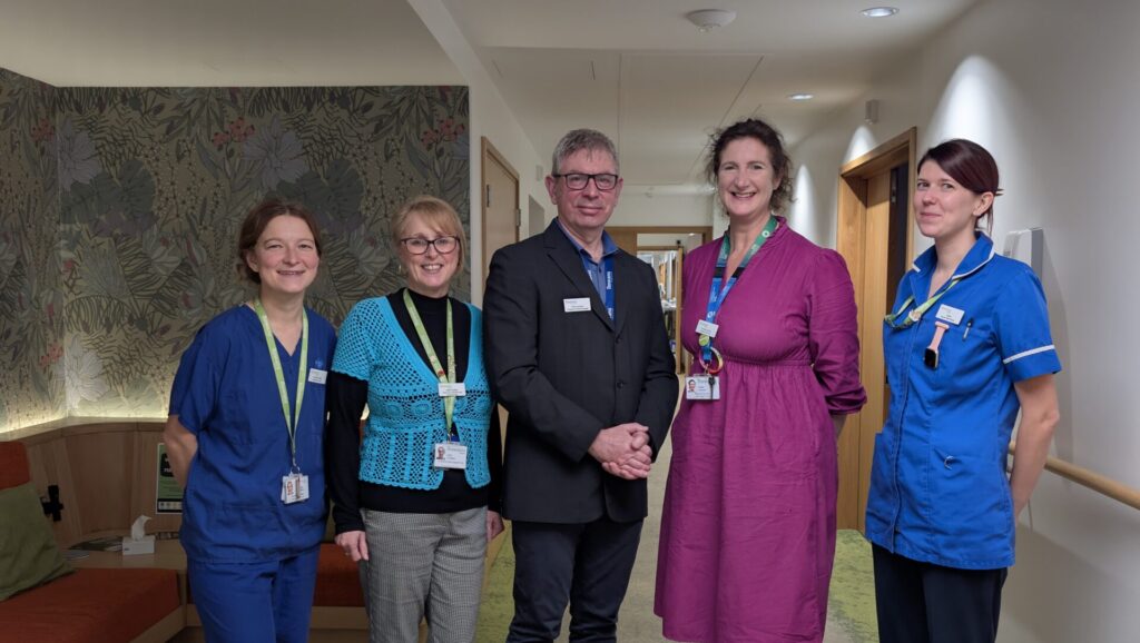 four woman and a man standing formally posing to camera. They all are smiling, and wearing green lanyards with ID badges. First woman on the left is wearing dark blue scrubs, to her right is a woman with short blond hair, glasses, black and light blue top and grey jeans. In the middle is a tall man with short hair, glasses, wearing a brown suit. To his right is a woman the same height as him wearing a pink dress. To her right is a woman, slightly shorter, wearing blue nursing scrubs.