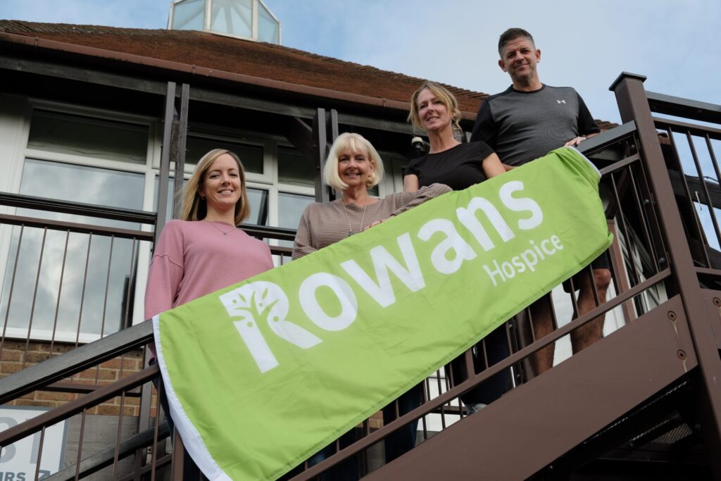 A group of four people—three women and one man—pose together on an outdoor staircase. They are holding a large, bright green banner that reads "Rowans Hospice" in white lettering, featuring a stylized tree logo. Image Overview The Group: The individuals are smiling and positioned at different levels on the stairs. The woman on the far left wears a pink long-sleeved shirt, while the others are dressed in casual tones like grey and black. The Setting: They are standing on a dark metal staircase attached to a brick building. In the background, there are large windows reflecting the sky and a distinctive glass cupola or skylight on the roof. The Banner: A lime-green rectangular banner is draped over the handrail, clearly displaying the "Rowans Hospice" branding.
