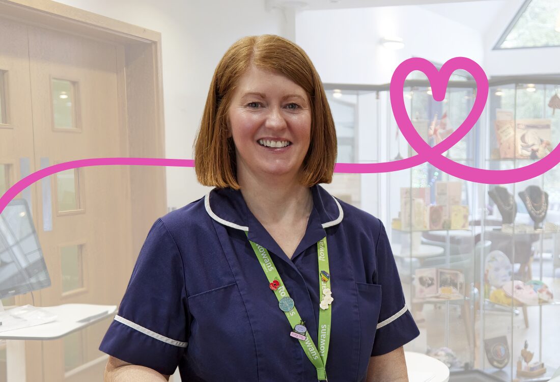 A smiling woman in a navy-blue nurse’s uniform stands indoors, facing the camera. She has shoulder-length ginger hair and wears a green lanyard with several pins attached. Behind her is a pink line going left to right transforming into a heart over her shoulder, the background is a bright, modern interior with glass display cabinets containing cards, crafts, and small gift items, as well as a wooden door to the left. The setting is a healthcare community space with a welcoming atmosphere.