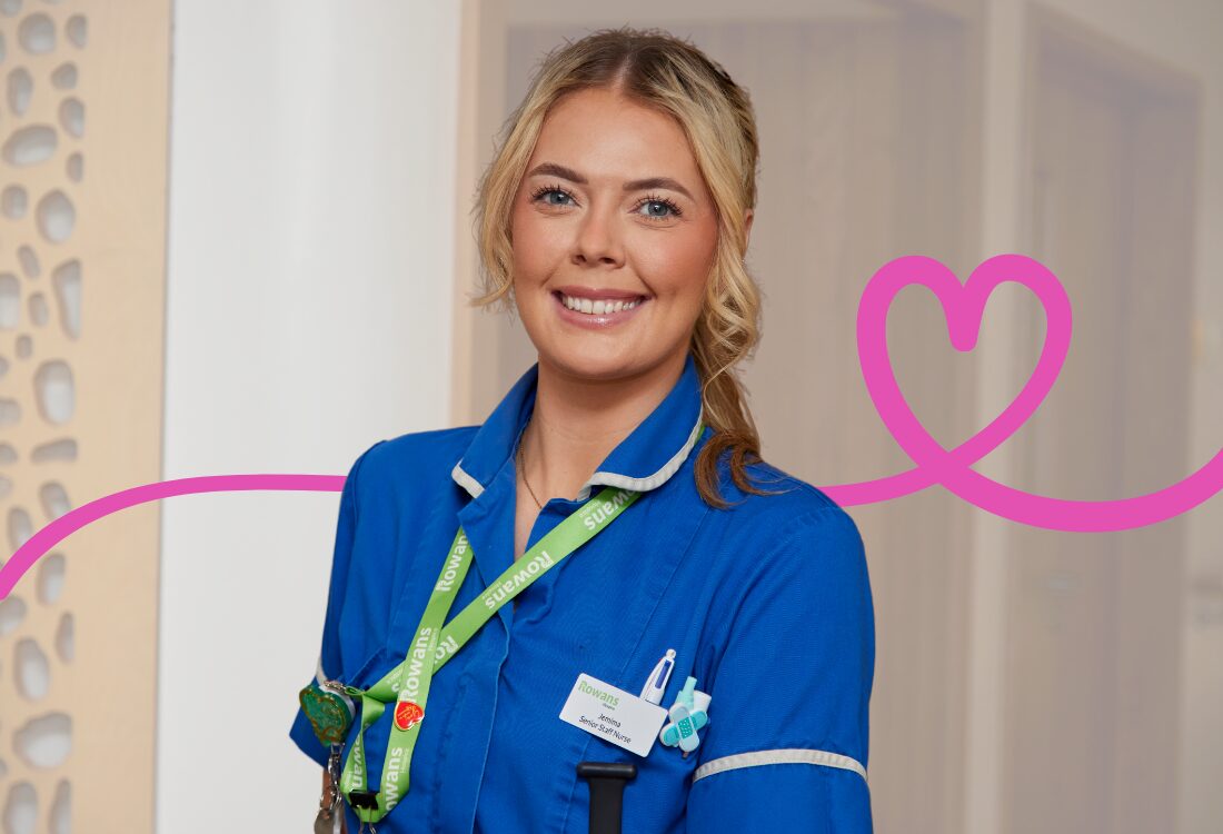A smiling woman in a blue nurse’s uniform stands indoors, facing the camera. She has blonde hair in a ponytails, and wears a green lanyard with several pins attached. Behind her is a pink line going left to right transforming into a heart over her shoulder, the background is a brown wooden wall which is out of focus with a transparent white layer.