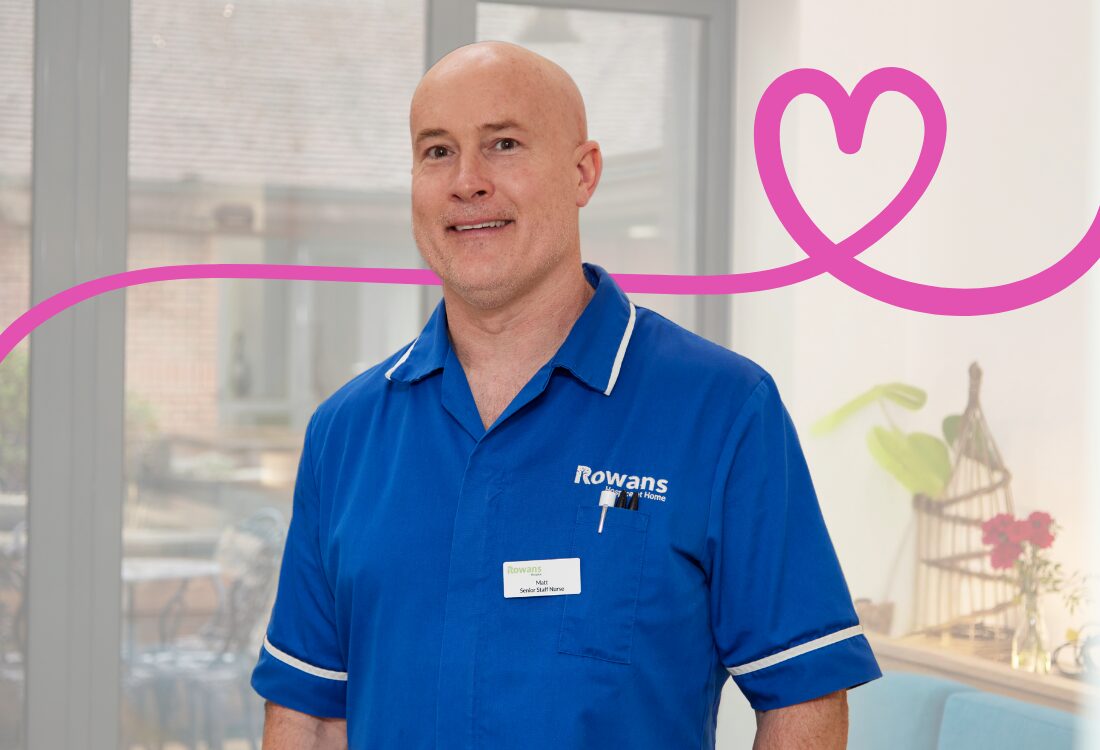 A smiling man in a blue nurse’s uniform stands indoors, facing the camera. He is bald has and wears a badge attached to the uniform top. Behind him is a pink line going left to right transforming into a heart over his shoulder. In the background, on his left, is a window through which you can see green grass and a building, on the right is a shelf with wooden decorations on.