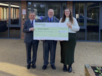 two men in formalwear holding a cheque presentation with a lady. The Man on the left is wearing a grey suit, white shirt, blue tie, black shoes, and smiles to the camera, he is shorter than the other two people, he has grey hair. The man in the middle is slightly taller, wears a grey suit with a blue tie and shirt. He also smiles to camera. The woman on the left is in her twenties, has long brown hair, she smiles to the camera, wearing a white knit jumper, a silk dark green skirt and black boots.