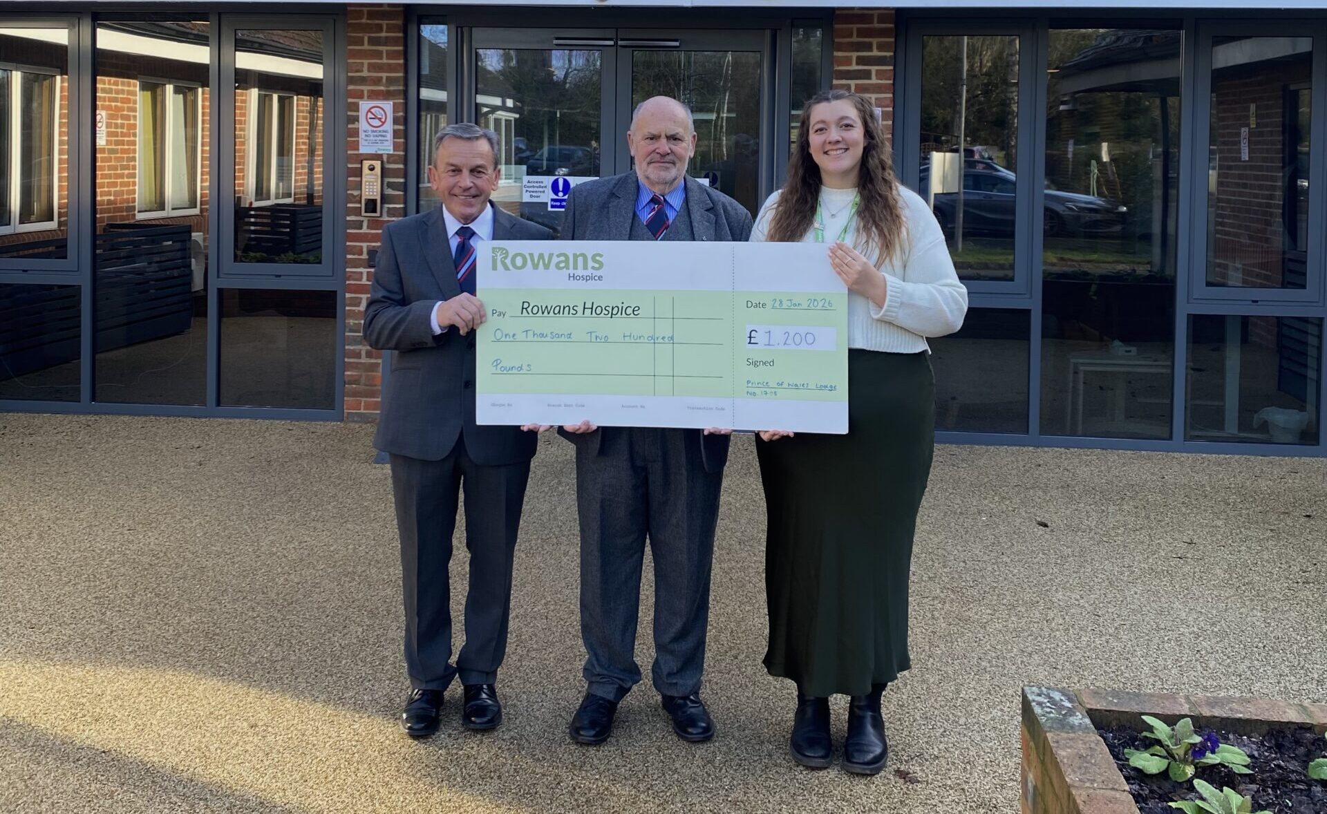 two men in formalwear holding a cheque presentation with a lady. The Man on the left is wearing a grey suit, white shirt, blue tie, black shoes, and smiles to the camera, he is shorter than the other two people, he has grey hair. The man in the middle is slightly taller, wears a grey suit with a blue tie and shirt. He also smiles to camera. The woman on the left is in her twenties, has long brown hair, she smiles to the camera, wearing a white knit jumper, a silk dark green skirt and black boots.