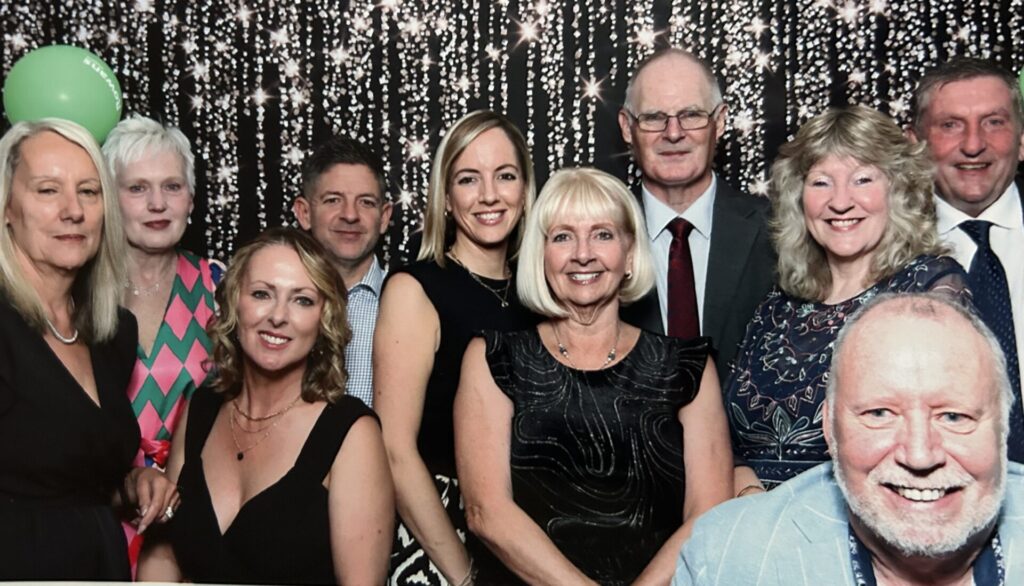 A medium shot captures a group of nine people posing for a photo against a shimmering, black-and-silver sequined backdrop. The Group and Attire Central Figures: In the front center, a woman with blonde hair and a black sleeveless dress smiles warmly. To her left, another woman in a black V-neck dress and gold necklaces leans in toward the center. Men in the Group: Behind the central woman stands a man in a dark suit with a red tie and glasses. To the far right, a man with a grey beard wearing a light blue blazer smiles prominently in the foreground. Another man in a grey suit and blue tie is visible in the upper right. Other Guests: On the far left, a woman in a black dress stands next to a woman wearing a vibrant pink and green patterned top. A green balloon is visible in the upper left corner. Setting and Composition Backdrop: The background consists of vertical strands of sparkling lights or sequins, creating a festive, "photo booth" style atmosphere. Lighting: The lighting is bright and even, highlighting the faces and festive clothing of the attendees. Mood: The overall tone is celebratory and formal.