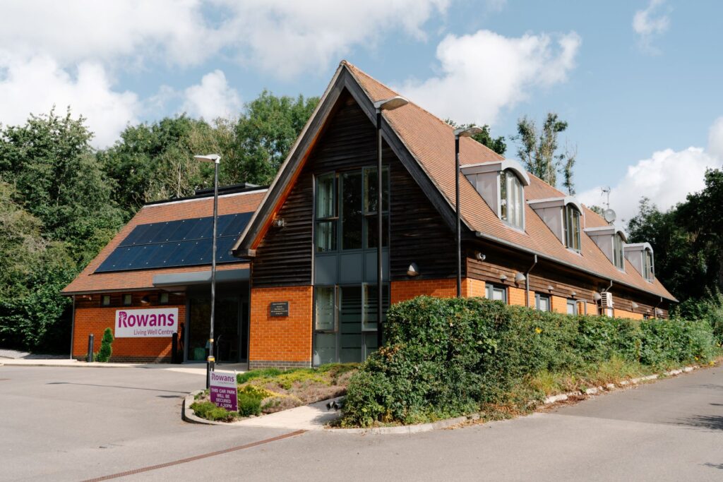 Brick Building with angled roof with solar panels. The build is in L shape and has a white sign with pink writting "Rowans Hospice Living Well Centre"