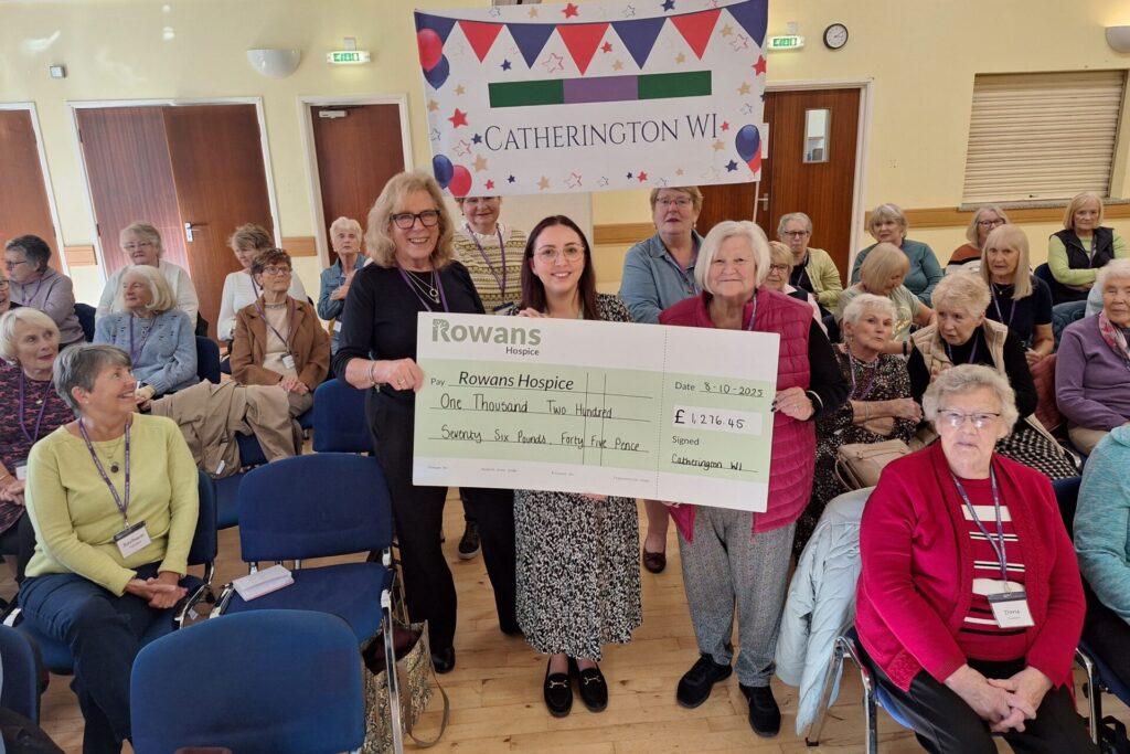 three women holding a giant check written to Rowans Hospice. The Woman on the left has blonde wavy hair, wears glasses and wears all black, she's in her early 70's/ The woman in the middle is in her mid 20's, she has round glasses, and dark red hair, she wears a long white and black dress. The woman on the right is older, has short white hair and wears a pink puffer gillet, a black long sleeve top underneath, grey trousers, black shoes.. Behind and above the women is a white banner that says "Catherington WI", in the room there are about 30 women sat on blue chairs. Some chairs are empty. The room looks like a village hall.