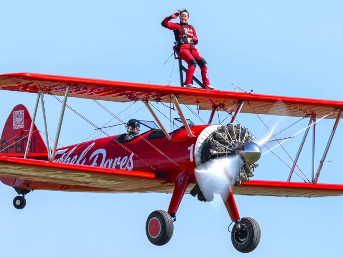 woman wearing a red boiler suit, on top of red biplane. The background is blue and looks like the plane is up in the sky