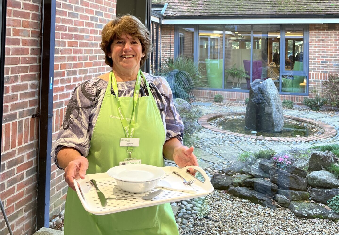 woman with short brown hair, smiling to camera and holding a tray. The woman is in her mid sixties, wearing a grey blouse and a green apron. She wears a green lanyard. On the tray is a bowl, a fork, a knife and a napkin. The woman stands in front of a window and behind her is a water fountain.