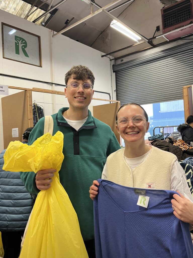 a lady and a man holding a yellow bag and a blue cardigan with a kilo sale of clothes on tables behind them