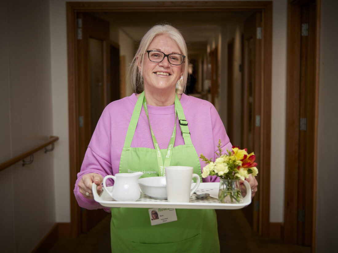 Woman with blond hair in a ponytail, wearing glasses, smiling to camera and holding a tray. She wears a pink jumper with a green apron over. on the tray is a white mug, a white jub and a small vase with bright yellow and red flowers. The woman stands in a corridor