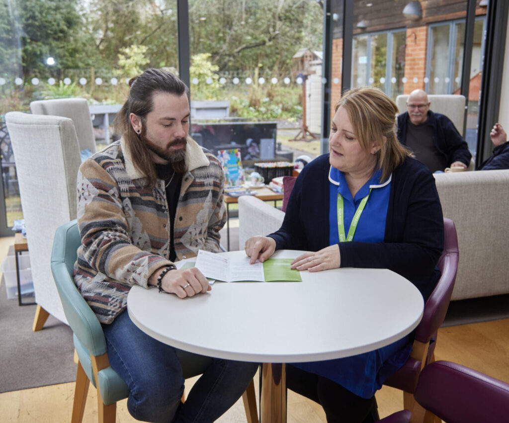 an image of one nurse sat at a white table talking to a man whilst reading a leaflet on the services with a garden behind them