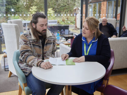 an image of one nurse sat at a white table talking to a man whilst reading a leaflet on the services with a garden behind them