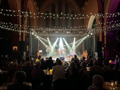 photo of a dimly lit room and stage at a concert with people in the view watching the stage