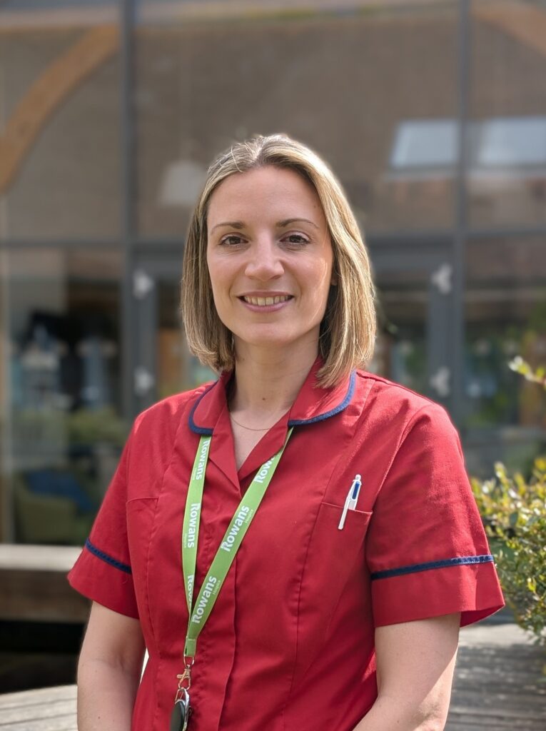 Woman in red nurses uniform smiling to camera. She has blond hair and brown eyes. The background is a glass building, blurred.