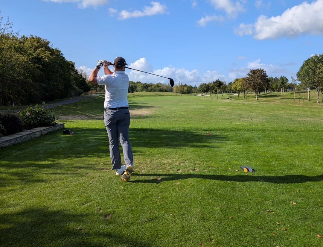 man with golf club after taking shot. The man has his back to the camera, wears a white polo shirt and grey trousers. He stands on a green filed, there's a blue sky with scattered clouds.