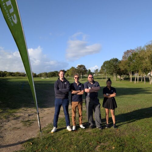 Three men, one woman, standing and looking to camera, holding golf clubs. Behind them is a flag with "Rowans Hospice" logo. They stand on a green field.