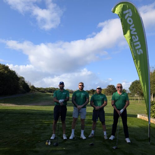 Four men standing and looking to camera, holding golf clubs. Behind them is a flag with "Rowans Hospice" logo. They stand on a green field. They all wear black shorts and green t-shirts.
