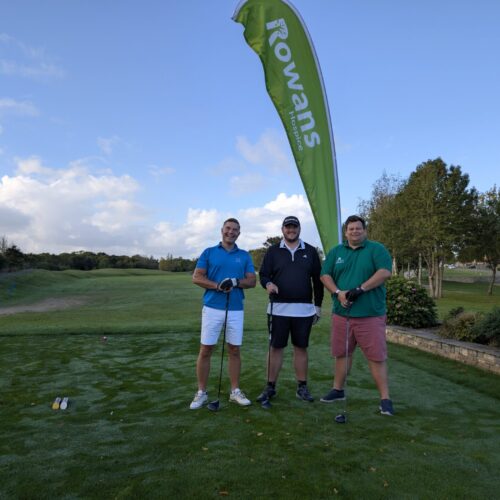 Three men standing and looking to camera, holding golf clubs. Behind them is a flag with "Rowans Hospice" logo. They stand on a green field.
