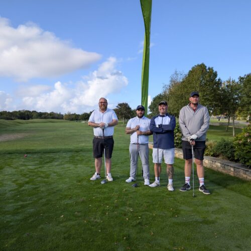 Four men standing and looking to camera, holding golf clubs. Behind them is a flag with "Rowans Hospice" logo. They stand on a green field. Three of them wear shorts, one wears torousers. All wear t-shirts.