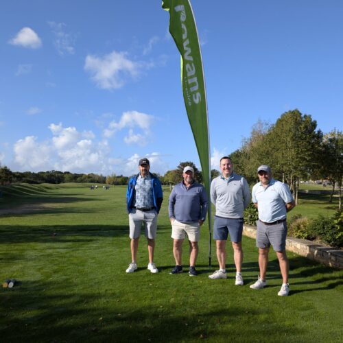 Four men standing and looking to camera, holding golf clubs. Behind them is a flag with "Rowans Hospice" logo. They stand on a green field. They all wear shorts and tshirts.