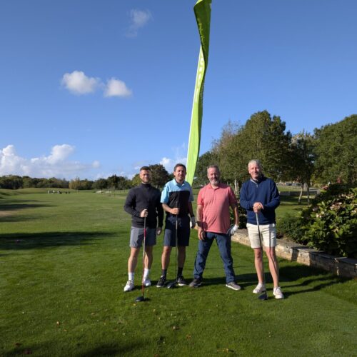 Four men standing and looking to camera, holding golf clubs. Behind them is a flag with "Rowans Hospice" logo. They stand on a green field.