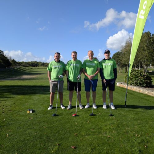 Four men standing and looking to camera, holding golf clubs. Behind them is a flag with "Rowans Hospice" logo. They stand on a green field. They all wear shorts and green t-shirts.