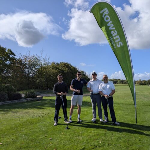 Four men standing and looking to camera, holding golf clubs. Behind them is a flag with "Rowans Hospice" logo. They stand on a green field.