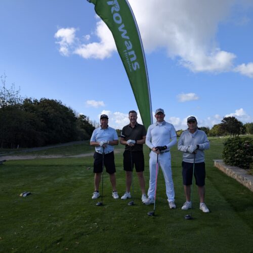 Four men standing and looking to camera, holding golf clubs. Behind them is a flag with "Rowans Hospice" logo. They stand on a green field.