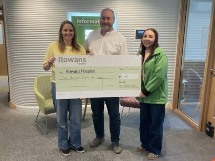 photo taken of three people in a reception area holding a cheque presentation fo over £2,000. The lady in green is from Rowans Hospice and the other two are a couple who have raised funds.