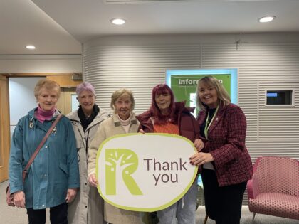 group photo in a waiting aeea with five people stood in a line holding a green thank you board with a Rowans R on