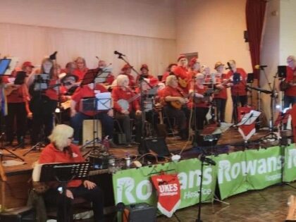 a large group of band members on a stage holding a green rowans hospice banner infront of them with instruments and microphones
