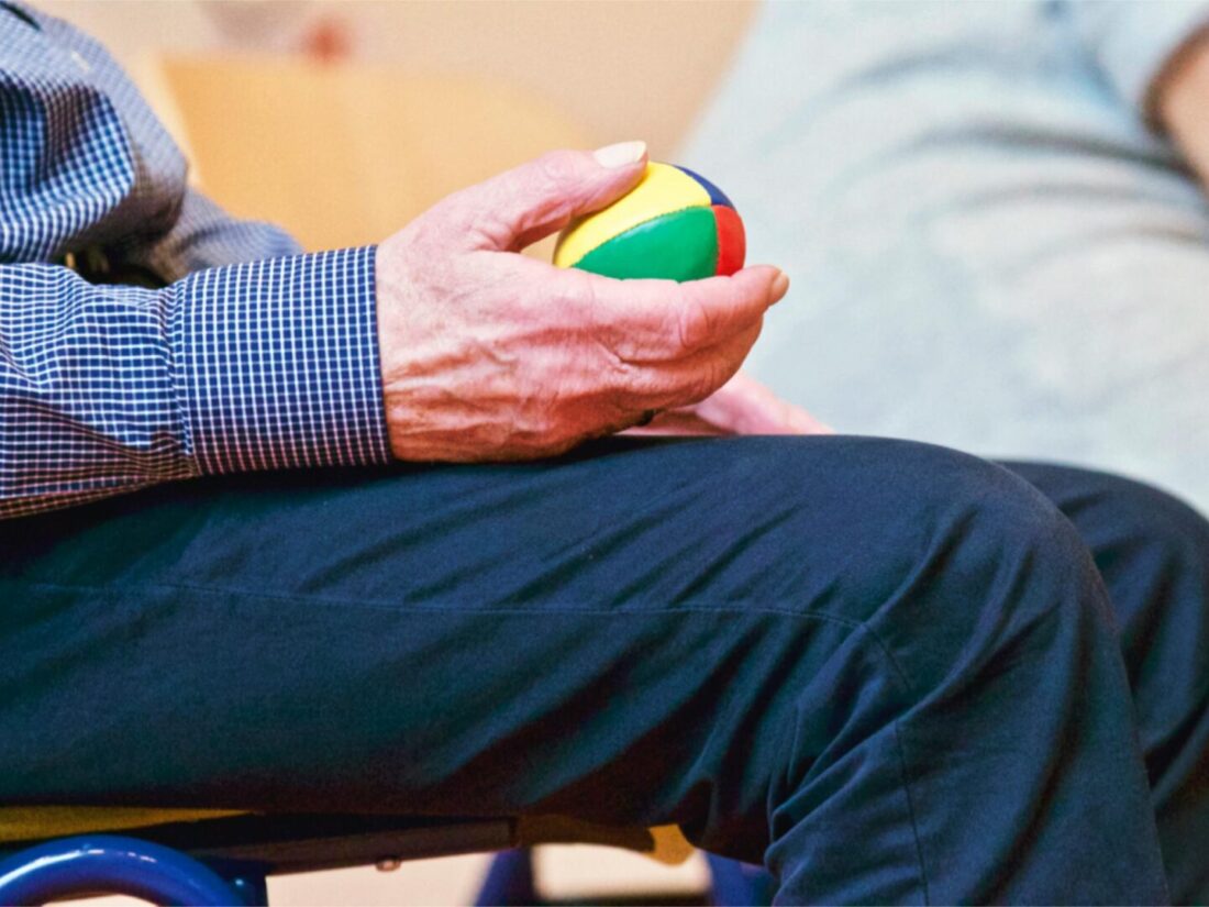 zoomed in photo of elderly man sat in a chair holding a colorful ball on his leg in a sports hall