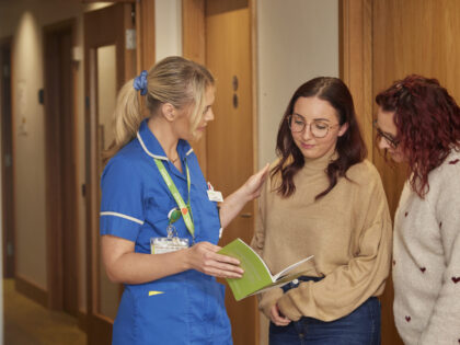 Woman with blond hair in a ponytail wearing blue nurse unifrom with had on another lady shoulder. This lady has dark red hair, glasses and wears a beige jumper, next to her is a woman with dark red wavy hair wearing a white jumper with red hearts. They all look at a leaflet.