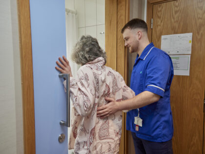 Man in blue nurse uniform holding the back of a slim woman with grey hair and in pink pyjamas. The woman has her right hand holding on to the door