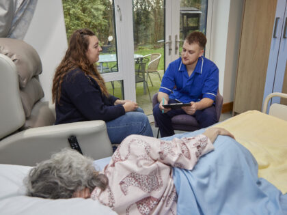 A healthcare professional in blue scrubs sits in a chair, holding a clipboard and speaking with a woman seated beside a hospital bed. In the foreground, an elderly patient lies on their side under a blanket, resting. The room is bright and calm, with a glass door opening onto a garden, suggesting a hospice or care setting focused on comfort and support.