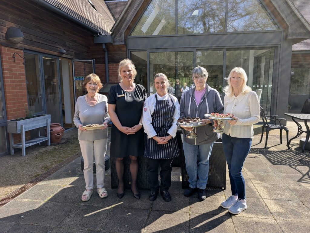 Group of five middle aged woman smiling to camera. The woman in the middle is dressed in chef's uniform, next to her the woman is blond and dressed in black. To her left stands a shorter woman holding a plate. On the other side, next to the chef, stand two women dressed in jeans and a casual top and holding a plate each.