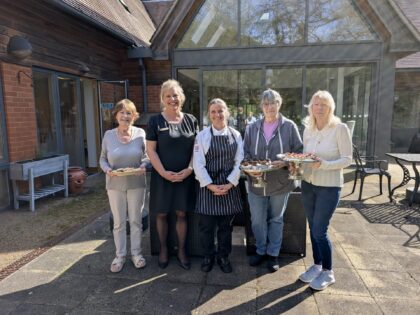 Group of five middle aged woman smiling to camera. The woman in the middle is dressed in chef's uniform, next to her the woman is blond and dressed in black. To her left stands a shorter woman holding a plate. On the other side, next to the chef, stand two women dressed in jeans and a casual top and holding a plate each.