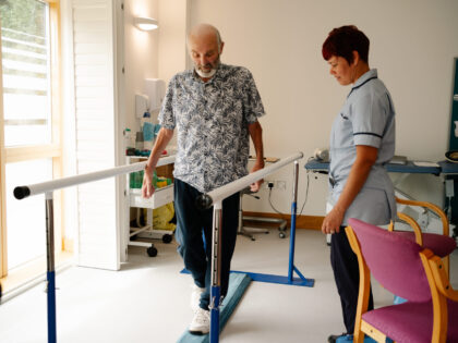 an older man with a beard diligently practices his mobility by walking on a balance beam between parallel bars while being closely monitored by a healthcare professional in a bright room.