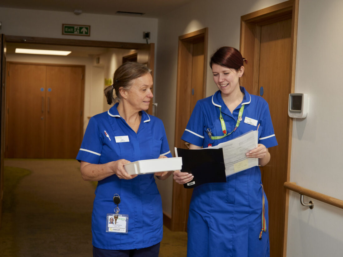 Two women walking down a corridor side by side. They are both nurses. The woman on the left has light brown hair in a ponytail and wears a blue scrubs top, she holds a white tray. The woman on the right is taller and wears a blue nurse unifrom dress, she holds a folder open. She has Dark red hair in an up do. They walk down a corridor with yellow dim lights.