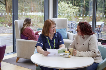 Two woman sat talking and smiling around a table. The woman on the left has ginger hair, shoulder length and straight, and wears a dark blue nurses uniform, the woman on the right has wavy dark red hair, she wears glasses and awhite knit jumper with small red hearts. They both smile and look at a brochure. 